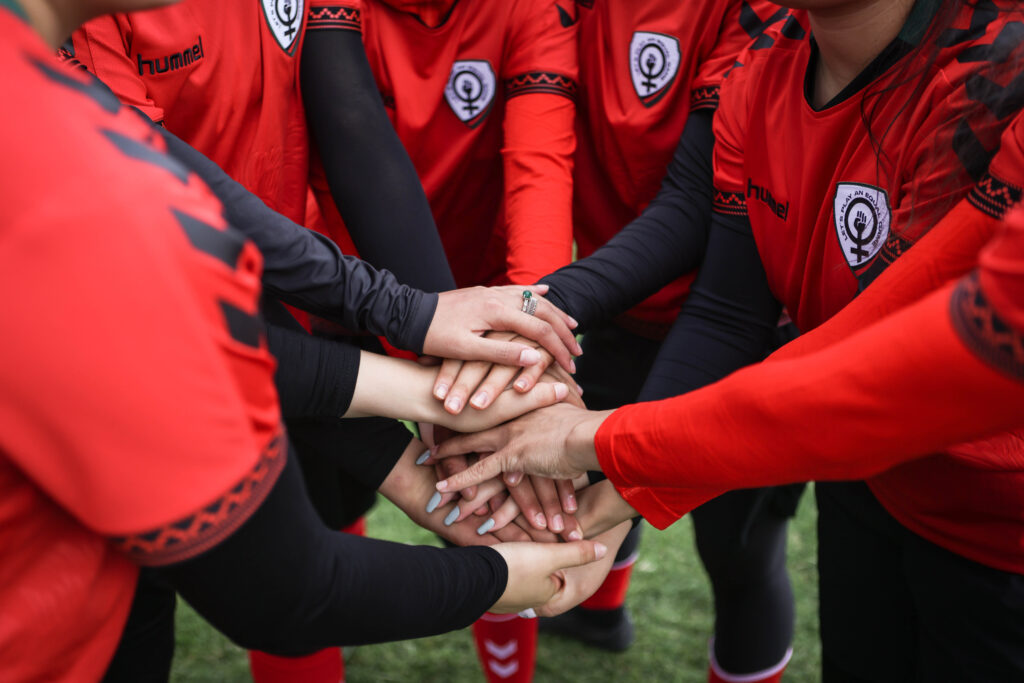 Image shows the arms of six players wearing read jerseys and black sleeves huddled with their hands together.